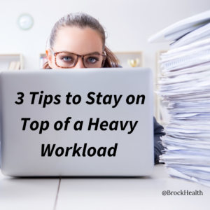 three tips to stay on top of a heavy workload. These words are written on the back of a laptop computer with a woman behind it and a stack of papers to the left